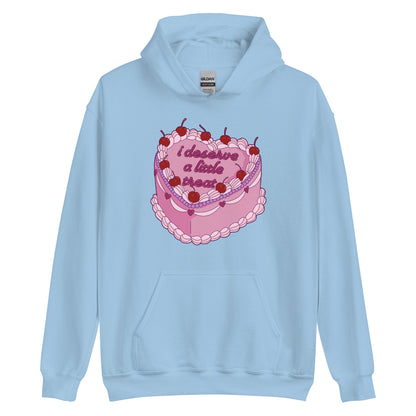 A light blue hooded sweatshirt featuring an illustration of an elaborately decorated pink cake. Icing on the cake reads "i deserve a little treat" in a cursive font.