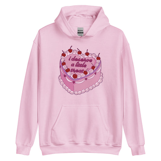 A light pink hooded sweatshirt featuring an illustration of an elaborately decorated pink cake. Icing on the cake reads "i deserve a little treat" in a cursive font.