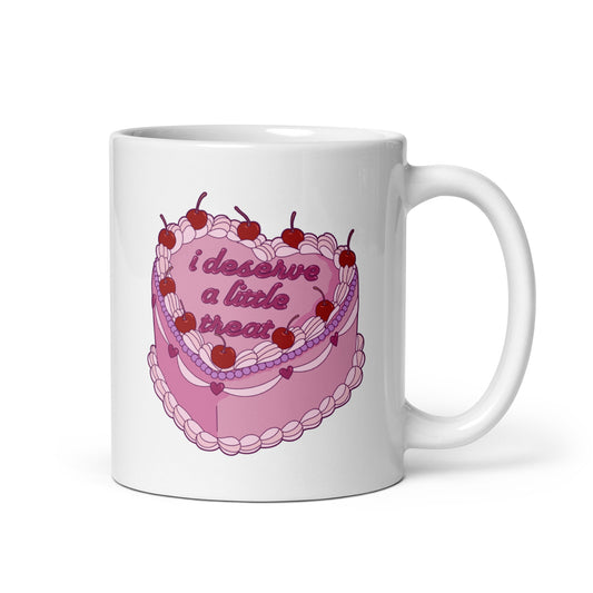An 11 ounce, white ceramic mug featuring an illustration of an elaborately decorated pink cake. Icing on the cake reads "i deserve a little treat" in a cursive font.