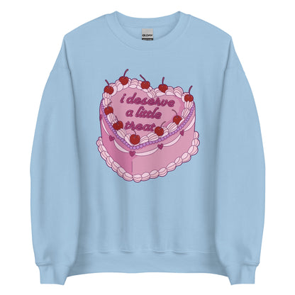 A light blue crewneck sweatshirt featuring an illustration of an elaborately decorated pink cake. Icing on the cake reads "i deserve a little treat" in a cursive font.