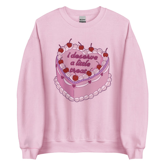 A light pink crewneck sweatshirt featuring an illustration of an elaborately decorated pink cake. Icing on the cake reads "i deserve a little treat" in a cursive font.