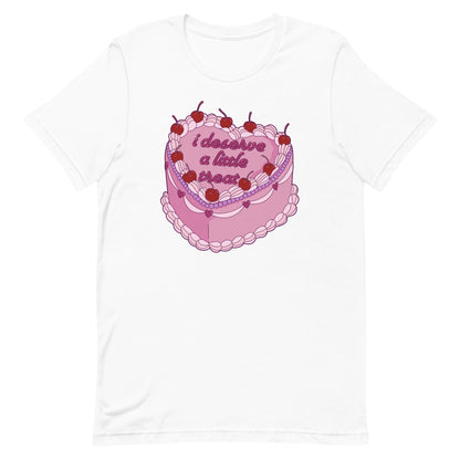 A white crewneck t-shirt featuring an illustration of an elaborately decorated pink cake. Icing on the cake reads "i deserve a little treat" in a cursive font.