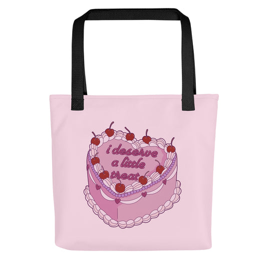 A pink tote bag with black handles, featuring an illustration of an elaborately decorated pink cake. Icing on the cake reads "i deserve a little treat" in a cursive font.