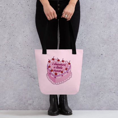 A waist-down image of a model wearing all black and standing against a concrete wall. The model is holding a pink tote bag with black handles, featuring an illustration of an elaborately decorated pink cake. Icing on the cake reads "i deserve a little treat" in a cursive font.