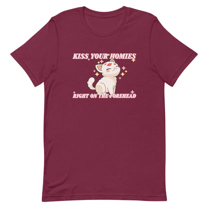 A maroon crewneck t-shirt featuring an illustration of a soft white kitten. Lipstick kiss marks are on the kitten's forehead, and its face is surrounded by sparkles. Pink text around the kitten reads "Kiss your homies right on the forehead."