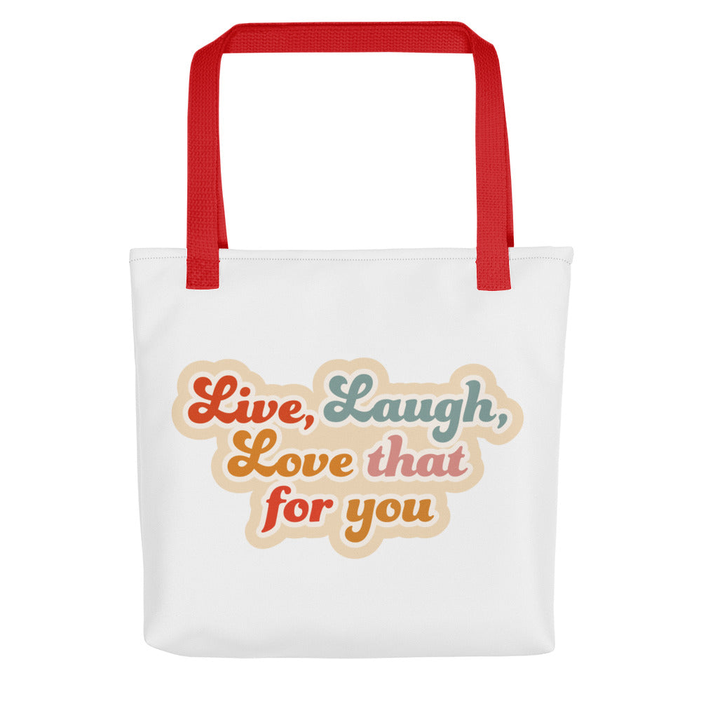 A white tote bag with red handles, featuring colorful, cursive text that reads "Live, Laugh, Love that for you"
