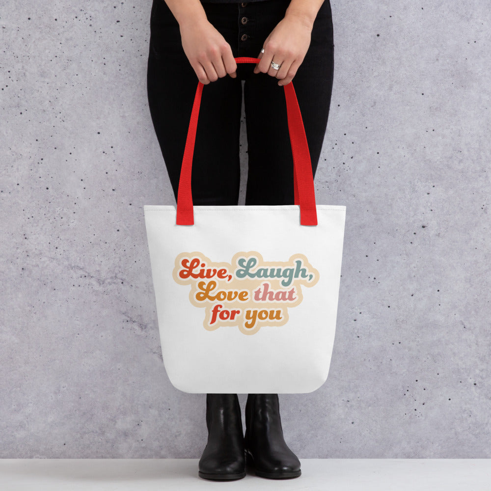 A waist-down image of a model wearing all black holding a white tote bag with red handles, featuring colorful, cursive text that reads "Live, Laugh, Love that for you"