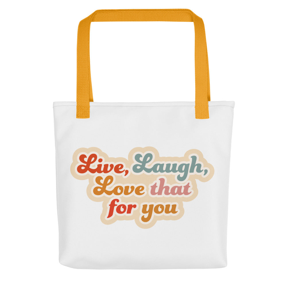A white tote bag with yellow handles, featuring colorful, cursive text that reads "Live, Laugh, Love that for you"