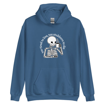 A indigo hooded sweatshirt featuring an illustration of a tired-looking skeleton holding a steaming mug. Text in an arc above the skeleton reads "too tired to live, too stubborn to die"