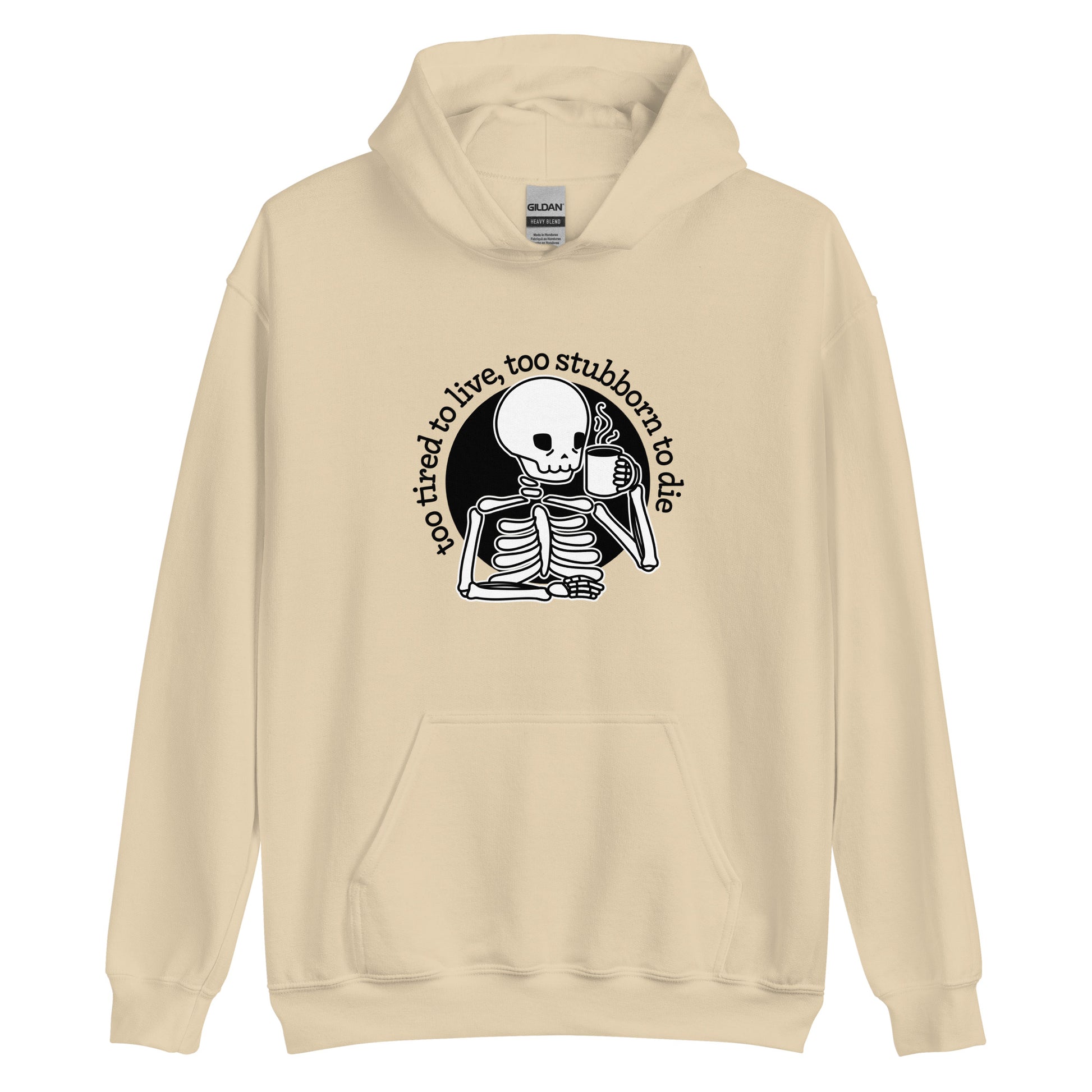 A cream-colored hooded sweatshirt featuring an illustration of a tired-looking skeleton holding a steaming mug. Text in an arc above the skeleton reads "too tired to live, too stubborn to die"