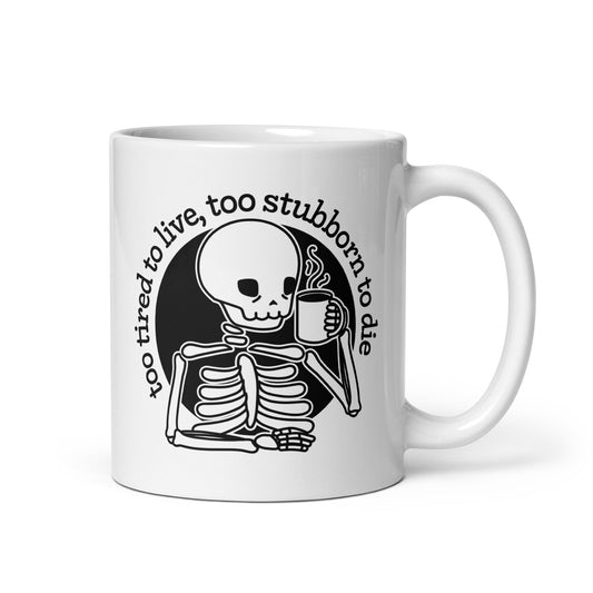 A white 11 ounce ceramic mug featuring a tired-looking skeleton holding a steaming mug. Text above the skeleton reads "too tired to live, too stubborn to die".