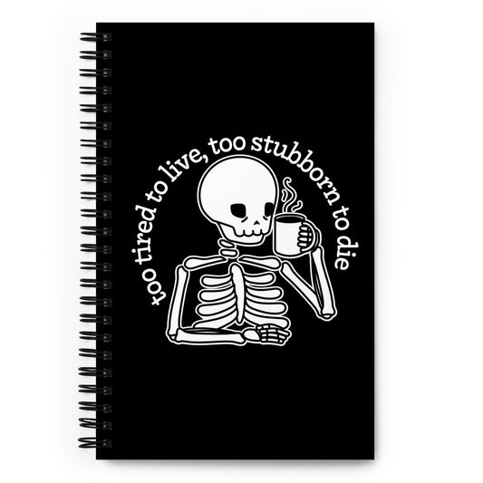 A black, wire-bound notebook featuring an illustration of a tired-looking skeleton holding a steaming mug. Text in an arc above the skeleton reads "too tired to live, too stubborn to die".