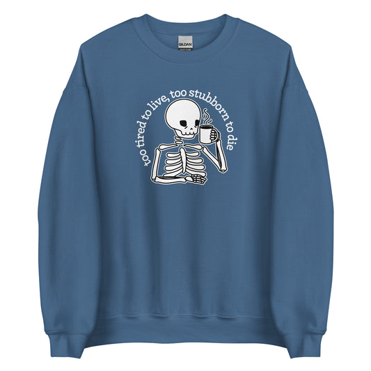 A blue crewneck sweatshirt featuring an illustration of a tired-looking skeleton holding a steaming mug. Text in an arc above the skeleton reads "too tired to live, too stubborn to die"