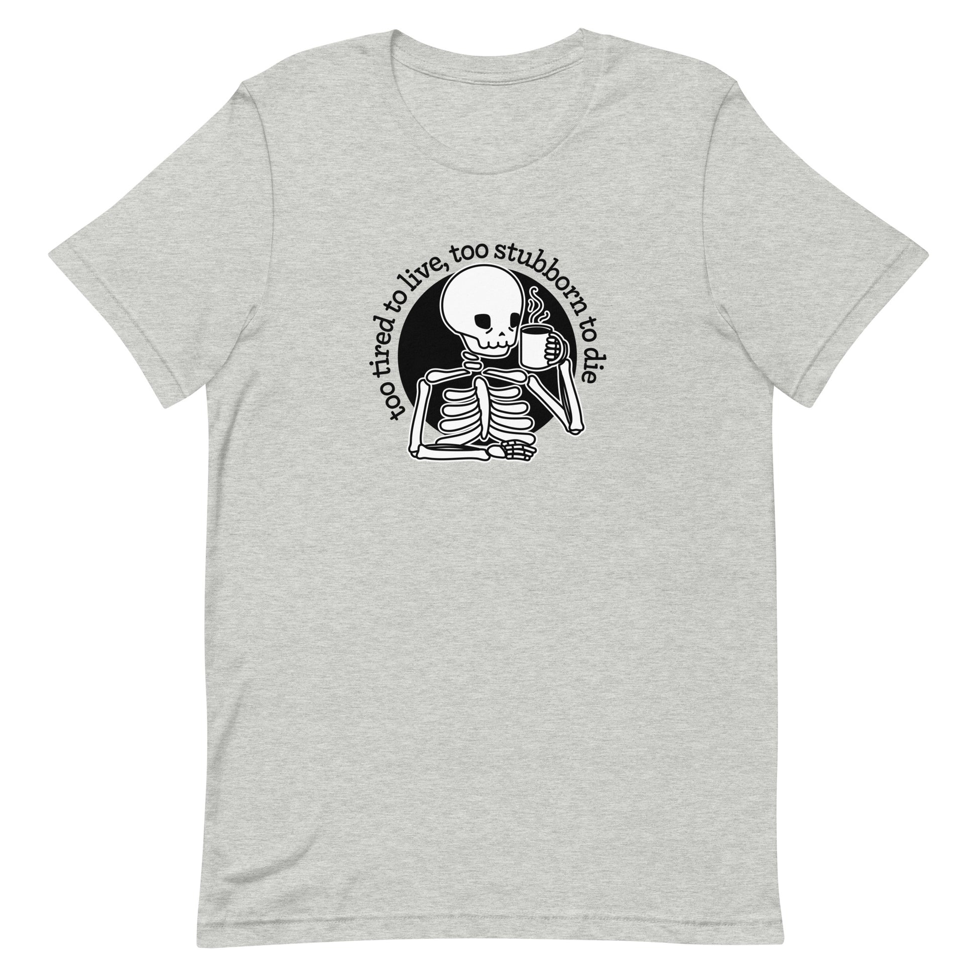 A grey crewneck t-shirt featuring a tired-looking skeleton holding a steaming mug. Text in an arc above the skeleton reads "too tired to live, too stubborn to die".