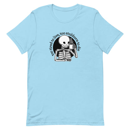 A light blue crewneck t-shirt featuring a tired-looking skeleton holding a steaming mug. Text in an arc above the skeleton reads "too tired to live, too stubborn to die".