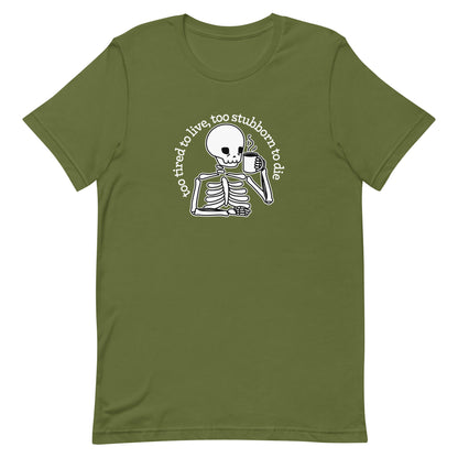 An olive green crewneck t-shirt featuring a tired-looking skeleton holding a steaming mug. Text in an arc above the skeleton reads "too tired to live, too stubborn to die".