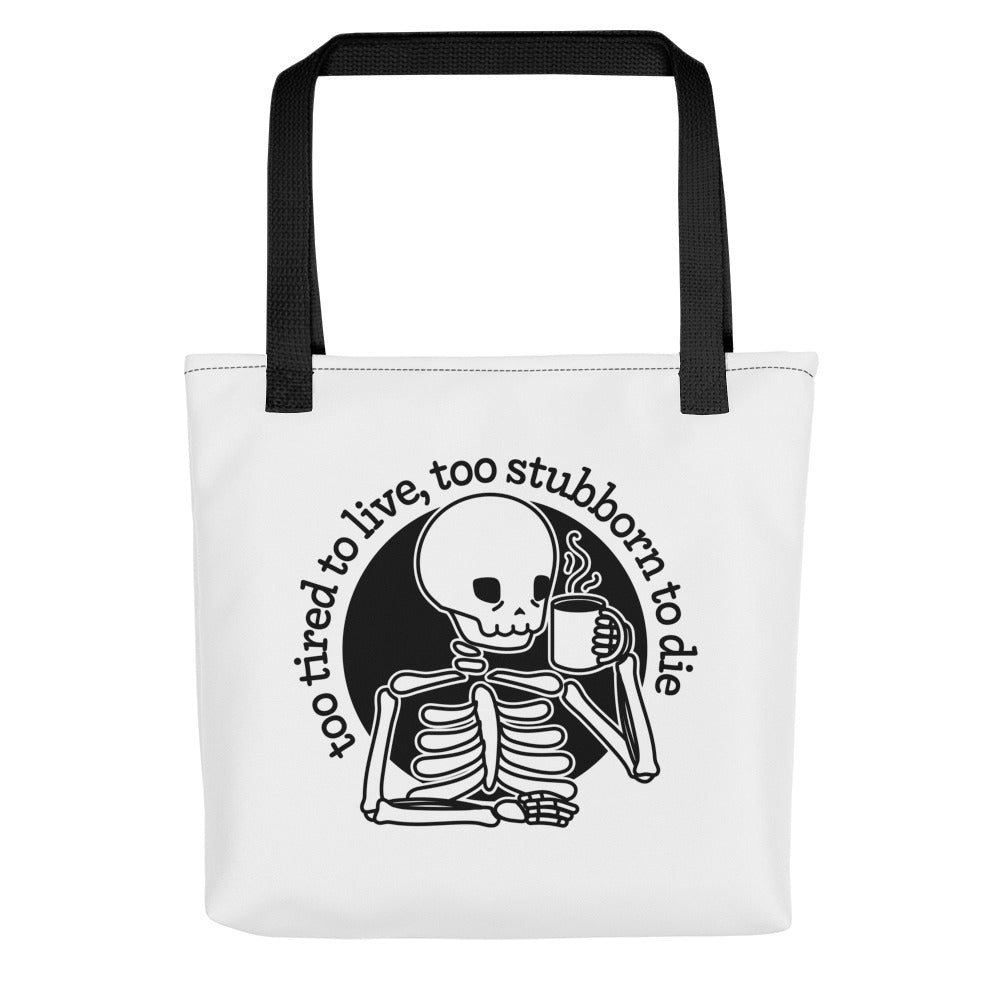 A white tote bag with black handles featuring an illustration of a tired skeleton holding a steaming mug. Text above the skeleton reads "too tired to live, too stubborn to die".