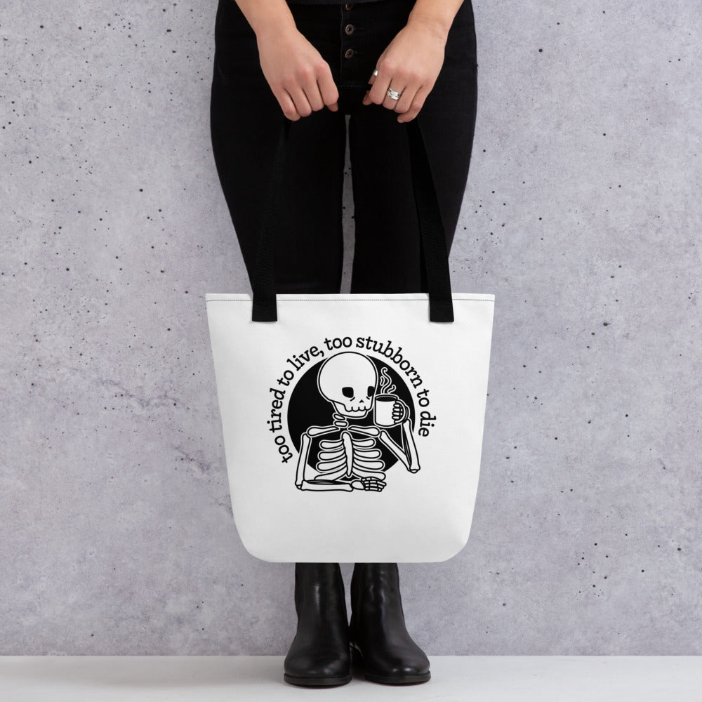 A waist-down image of a model wearing all black, holding a white tote bag with black handles. The bag features an illustration of a tired skeleton holding a steaming mug. Text above the skeleton reads "too tired to live, too stubborn to die".
