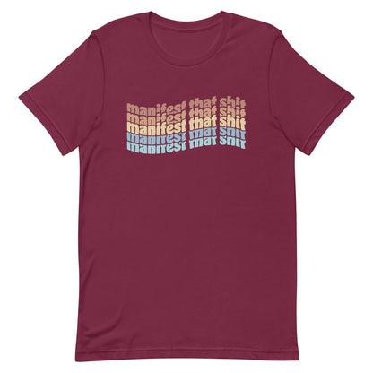 A maroon crewneck t-shirt featuring stacked text that reads "manifest that shit" in a rainbow of colors.