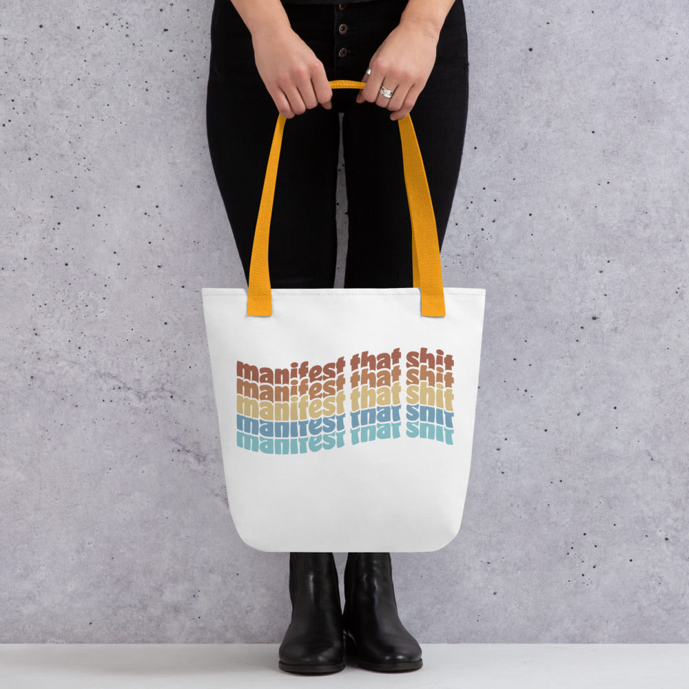 A waist-down image of a model wearing all black and holding a white tote bag with yellow handles featuring stacked text that reads "manifest that shit" in a rainbow of colors.