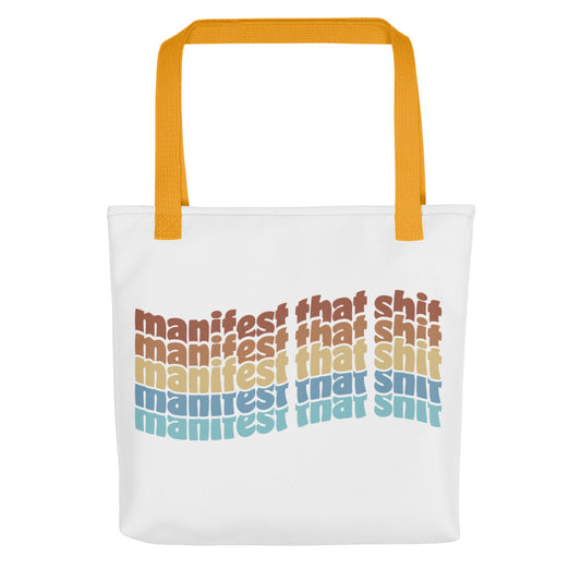 A white tote bag with yellow handles featuring stacked text that reads "manifest that shit" in a rainbow of colors.