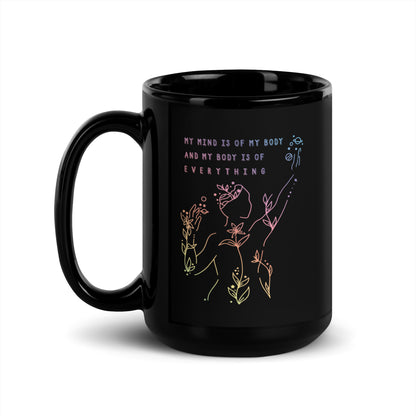 A black 15 ounce coffee mug featuring an abstract illustration of a figure whose body is spreading out into plants and planets and stars. Text above the figure reads "My mind is of my body and my body is of everything."
