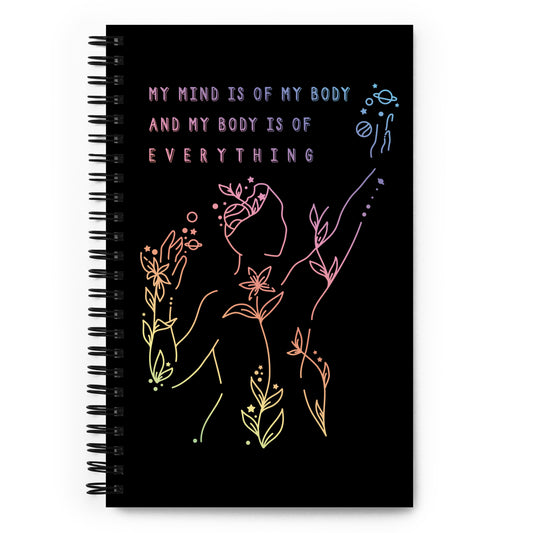 A black wire-bound notebook. The notebook features an abstract illustration of a figure whose body is spreading out into plants and planets and stars. Text above the figure reads "My mind is of my body and my body is of everything."