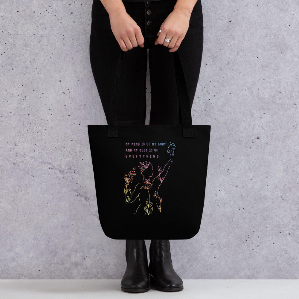 A waist-down image of a model wearing all black and holding a black canvas tote bag with a black handle. The tote bag features an abstract illustration of a figure whose body is spreading out into plants and planets and stars. Text above the figure reads "My mind is of my body and my body is of everything."