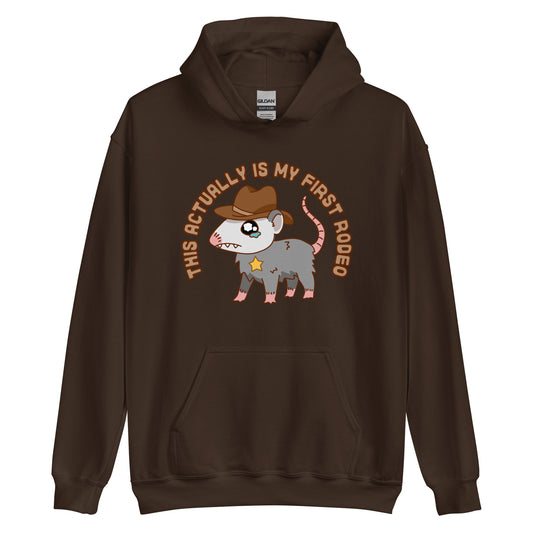 A brown hooded sweatshirt featuring an illustration of a cute and nervous possum wearing a cowboy hat and sherrif's star badge. Text above the possum in an arc reads "this actually is my first rodeo".