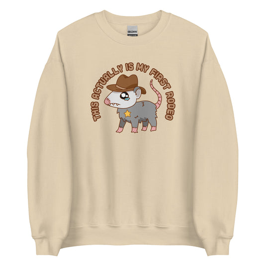 A tan crewneck sweatshirt featuring an illustration of a cute and nervous possum wearing a cowboy hat and sherrif's star badge. Text above the possum in an arc reads "this actually is my first rodeo".