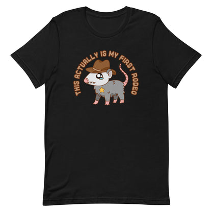 A black crewneck t-shirt featuring an illustration of a cute and nervous possum wearing a cowboy hat and sherrif's star badge. Text above the possum in an arc reads "this actually is my first rodeo".