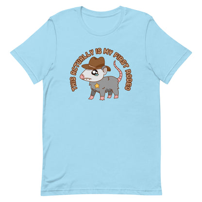A blue crewneck t-shirt featuring an illustration of a cute and nervous possum wearing a cowboy hat and sherrif's star badge. Text above the possum in an arc reads "this actually is my first rodeo".