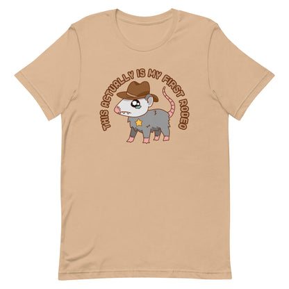 A tan crewneck t-shirt featuring an illustration of a cute and nervous possum wearing a cowboy hat and sherrif's star badge. Text above the possum in an arc reads "this actually is my first rodeo".