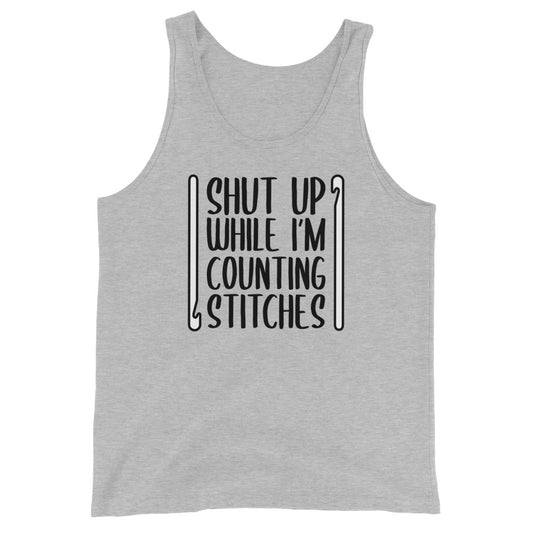 A grey tank top featuring black text that reads "Shut up while I'm counting stitches." The text is framed by a crochet hook to the left and right.