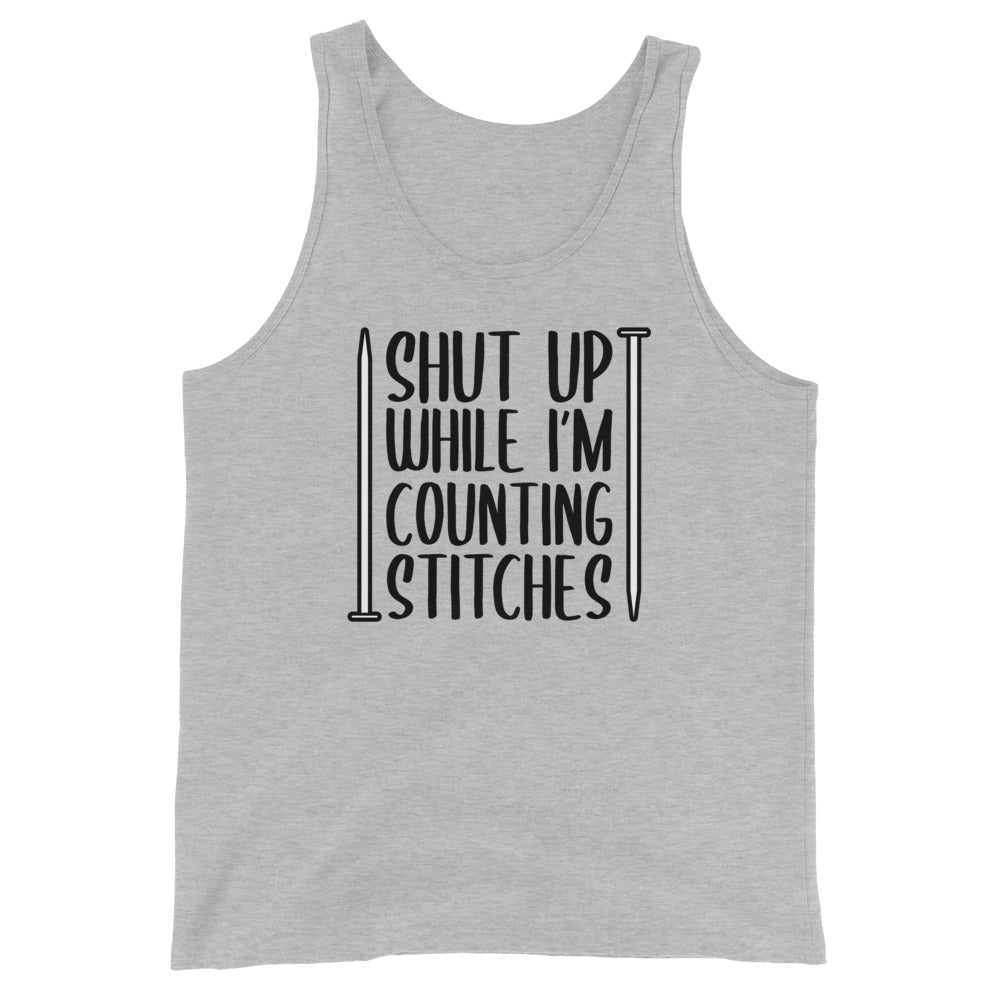 A grey tank top with black text surrounded by two knitting needles. The text reads "Shut up while I'm counting stiches".