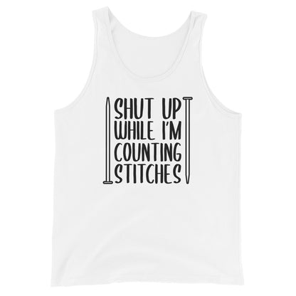 A white tank top with black text surrounded by two knitting needles. The text reads "Shut up while I'm counting stiches".