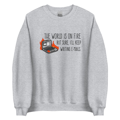 A light grey crewneck sweatshirt featuring a squiggly illustration of a laptop that is on fire. Text alongside the laptop reads "the world is on fire but sure, I'll keep writing e-mails."