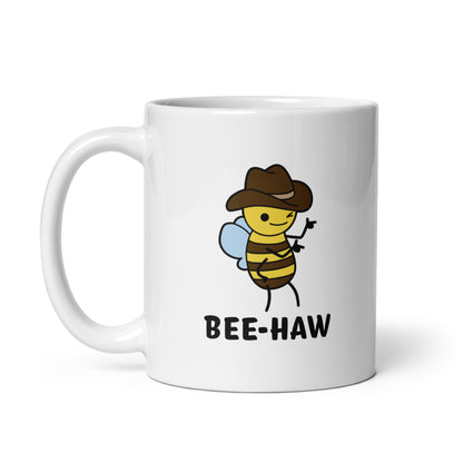 An 11 ounce white ceramic mug with an image of a bee in a cowboy hat on it. The bee is winking and holding up "finger guns". Text beneath the bee reads "Bee-Haw"