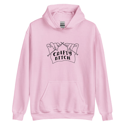 A light pink hooded sweatshirt featuring a single-color illustration of a pair of hands holding knitting needles. Fabric on the needles features text that reads "crafty bitch".