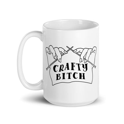 A white ceramic 15 ounce coffee mug featuring a single-color illustration of a pair of hands holding knitting needles. Fabric on the needles features text that reads "crafty bitch".