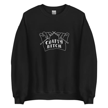 A black crewneck sweatshirt featuring a single-color illustration of a pair of hands holding knitting needles. Fabric on the needles features text that reads "crafty bitch".