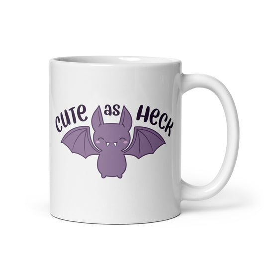 An 11 ounce white ceramic mug featuring an illustration of a smiling purple bat. Above the bat is text that reads "Cute As Heck"