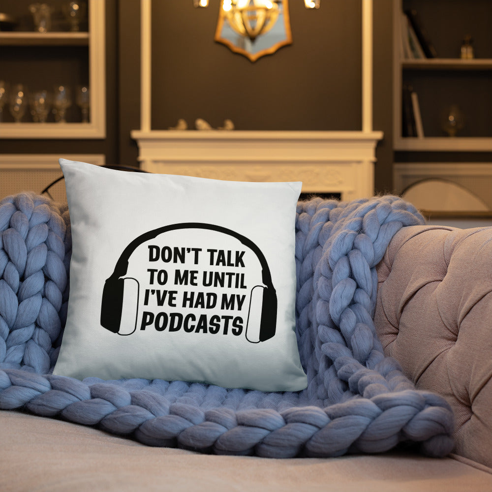 A white throw pillow with an image of headphones surrounding text that reads "Don't talk to me until I've had my podcasts." The pillow is resting on a gray couch with a blue blanket.