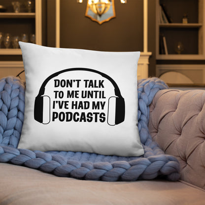 A white throw pillow with an image of headphones surrounding text that reads "Don't talk to me until I've had my podcasts." The pillow is resting on a gray couch with a blue blanket.
