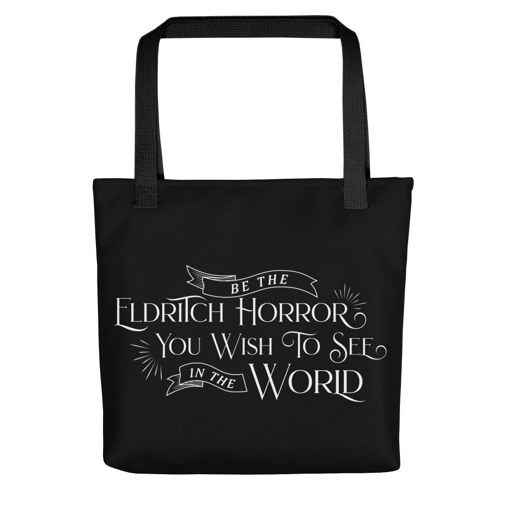 A solid black totebag featuring white text in an old-fashioned style that reads "Be the Eldritch Horror You Wish To See In The World"