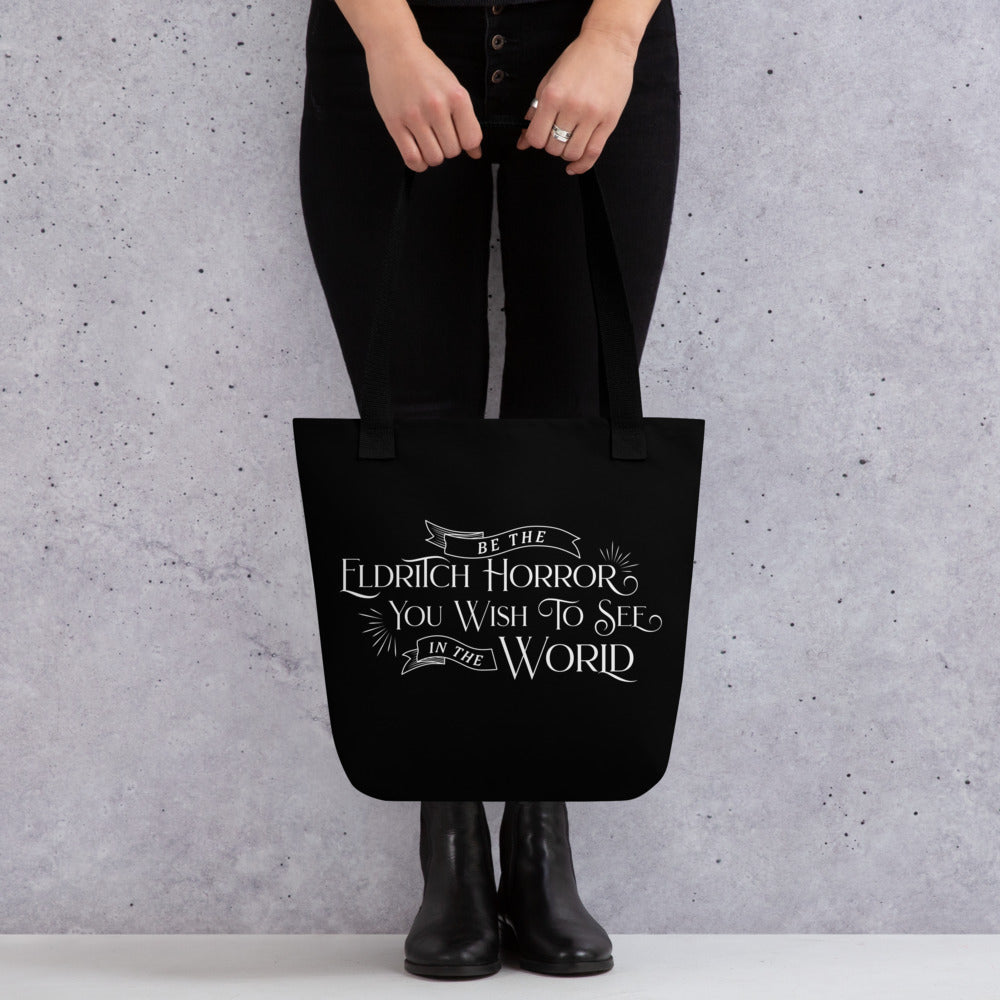 A waist-down image of a person standing against a concrete wall. The person is holding a black tote bag featuring text in an old-fashioned style that reads "Be The Eldritch Horror You Wish To See In The World"