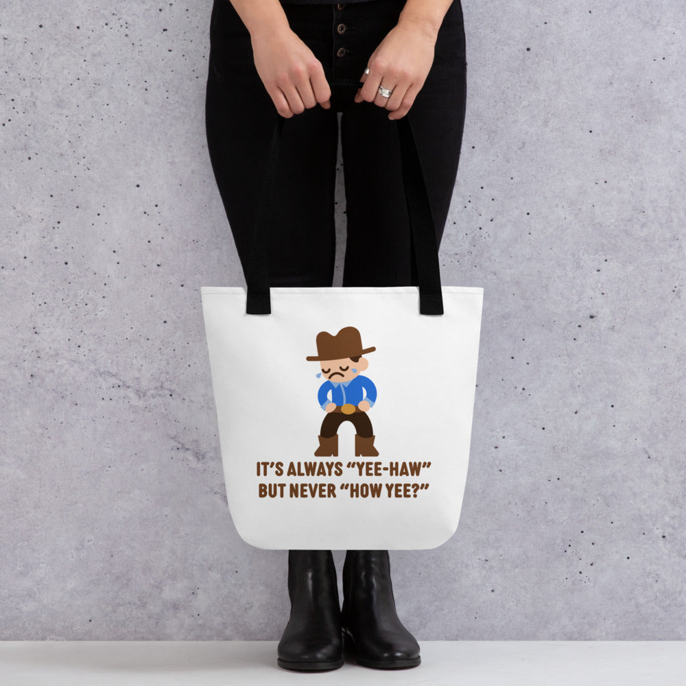 A waist-down image of a model wearing all black. The model is holding a white canvas tote bag with black handles featuring an illustration of a crying cowboy wearing a blue shirt. Text alongside the cowboy reads "It's always "yee-haw" but never "How yee?""