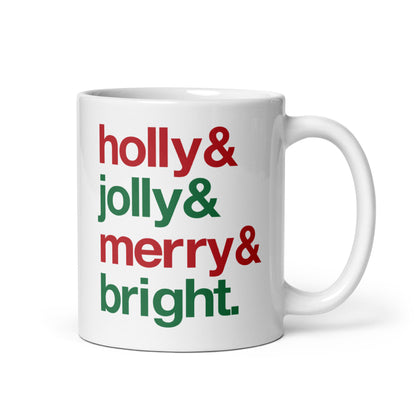 A white 11 ounce ceramic mug with four lines of red and green text that read "Holly & Jolly & Merry & Bright"