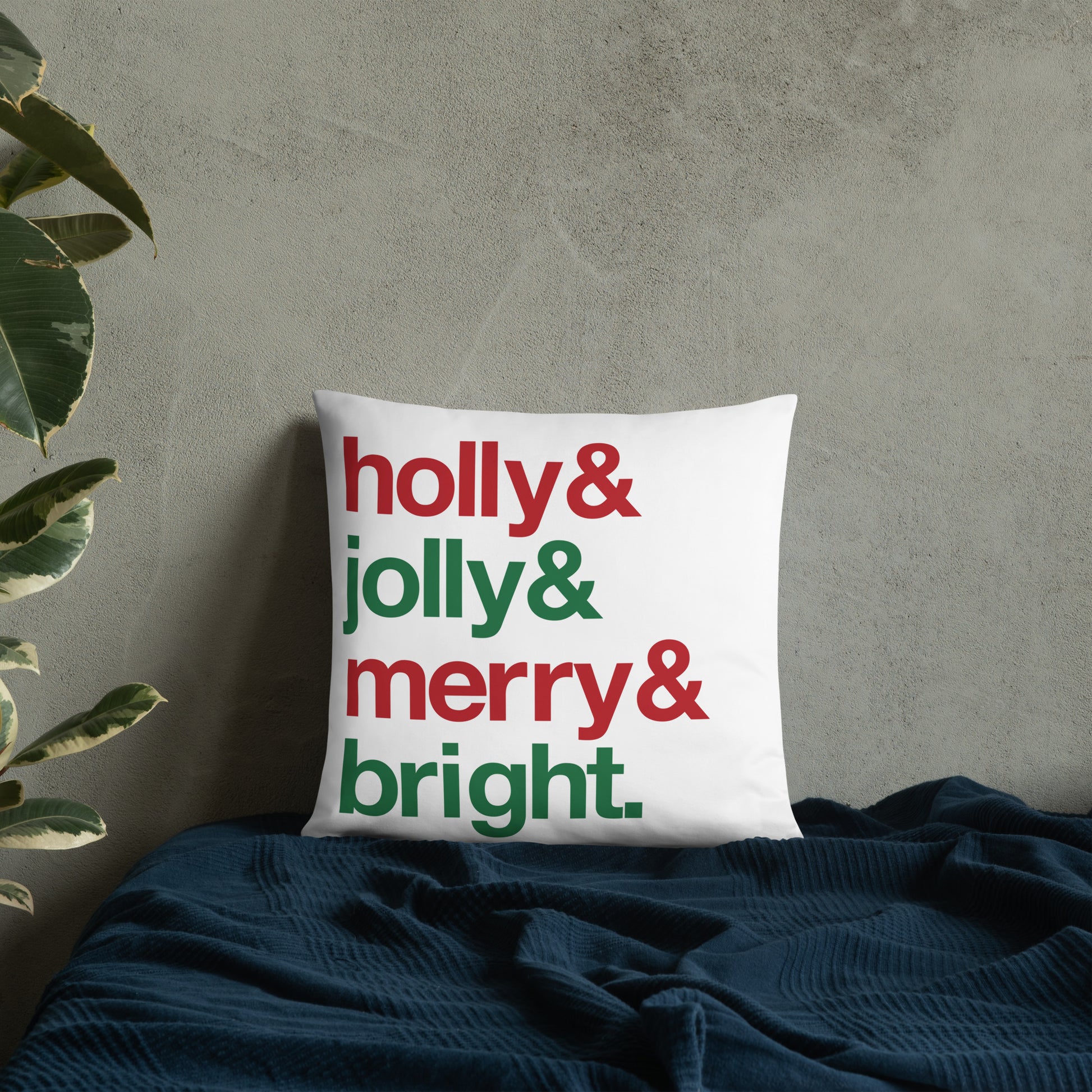 An 18" by 18" throw pillow decorated with four lines of red and green text that read "Holly & jolly & merry & bright". The pillow is  set on top of a bed with a blue blanket against a concrete wall.