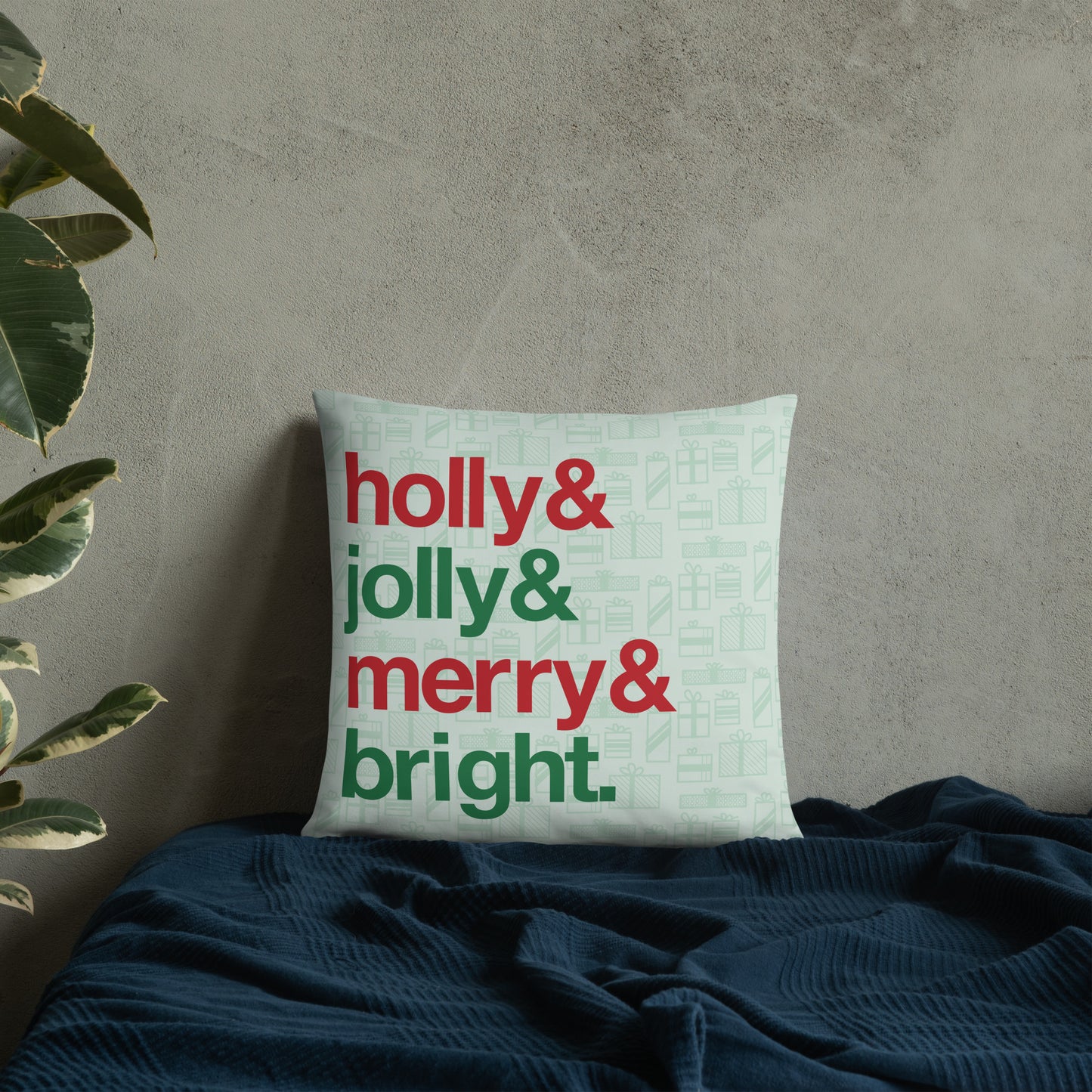 An 18" by 18" throw pillow decorated with a pale green background pattern of presents. There are also four lines of red and green text on the pillow that read "Holly & jolly & merry & bright". The pillow is  set on top of a bed with a blue blanket against a concrete wall.